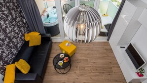 En moderne stue med en sort sofa, gule puder, et rundt sofabord med snacks, en usædvanlig hængende lampe og udsigt til et spabad og udendørs siddeområde.