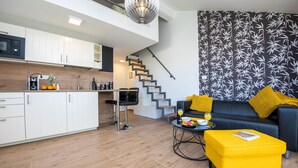En moderne lejlighed med et køkken, spiseområde og opholdsstue. Den har en trappe, en sort lædersofa med gule puder og en klar gul skammel.