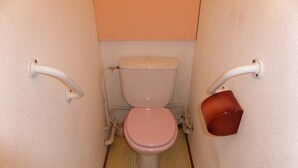 A small bathroom with a toilet, a pink seat, and a red bucket on the wall.