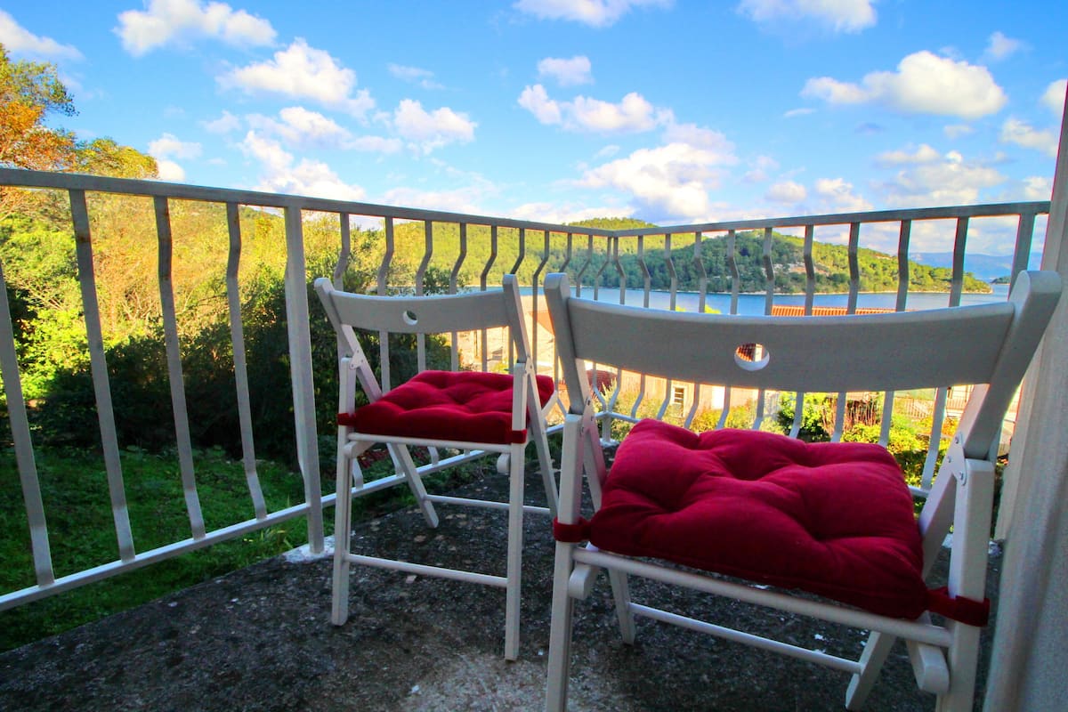 Two white chairs with red cushions on a balcony with a metal railing, overlooking a body of water and greenery.