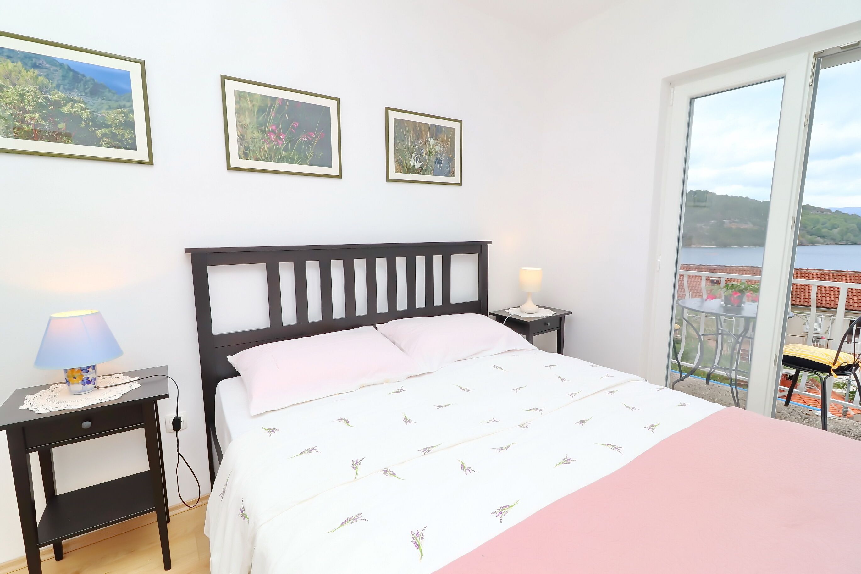 A bedroom with a bed, a nightstand with a lamp, and a view of a balcony with a table and chair.