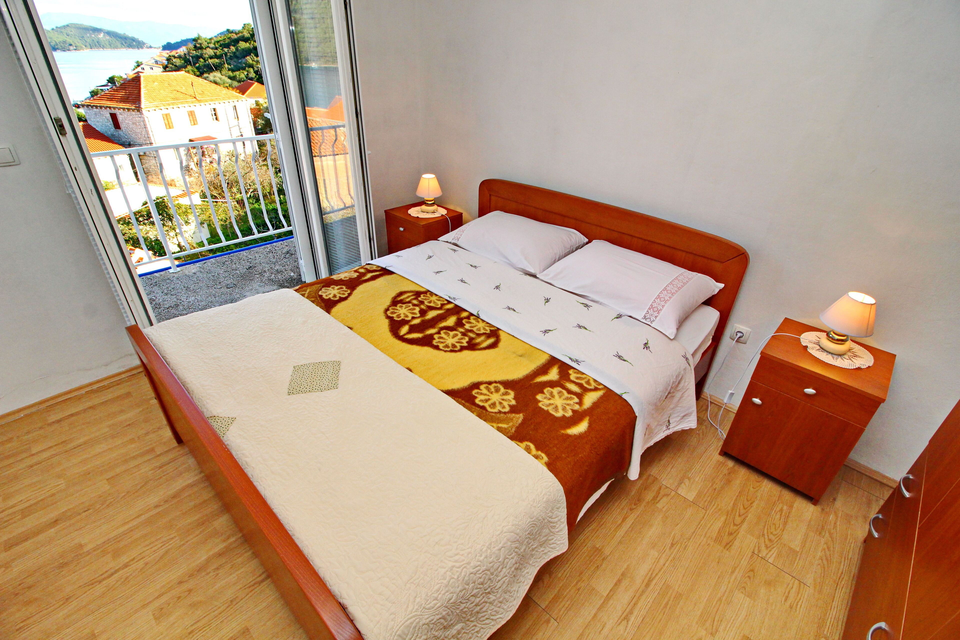 Ein Schlafzimmer mit einem Bett, zwei Nachttischen, Holzboden und Blick auf ein Gebäude und die Landschaft vom Balkon aus.