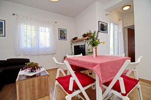 A dining area with a table covered in a red tablecloth, white chairs with red cushions, a small side table with a vase of flowers, and a television on a stand.