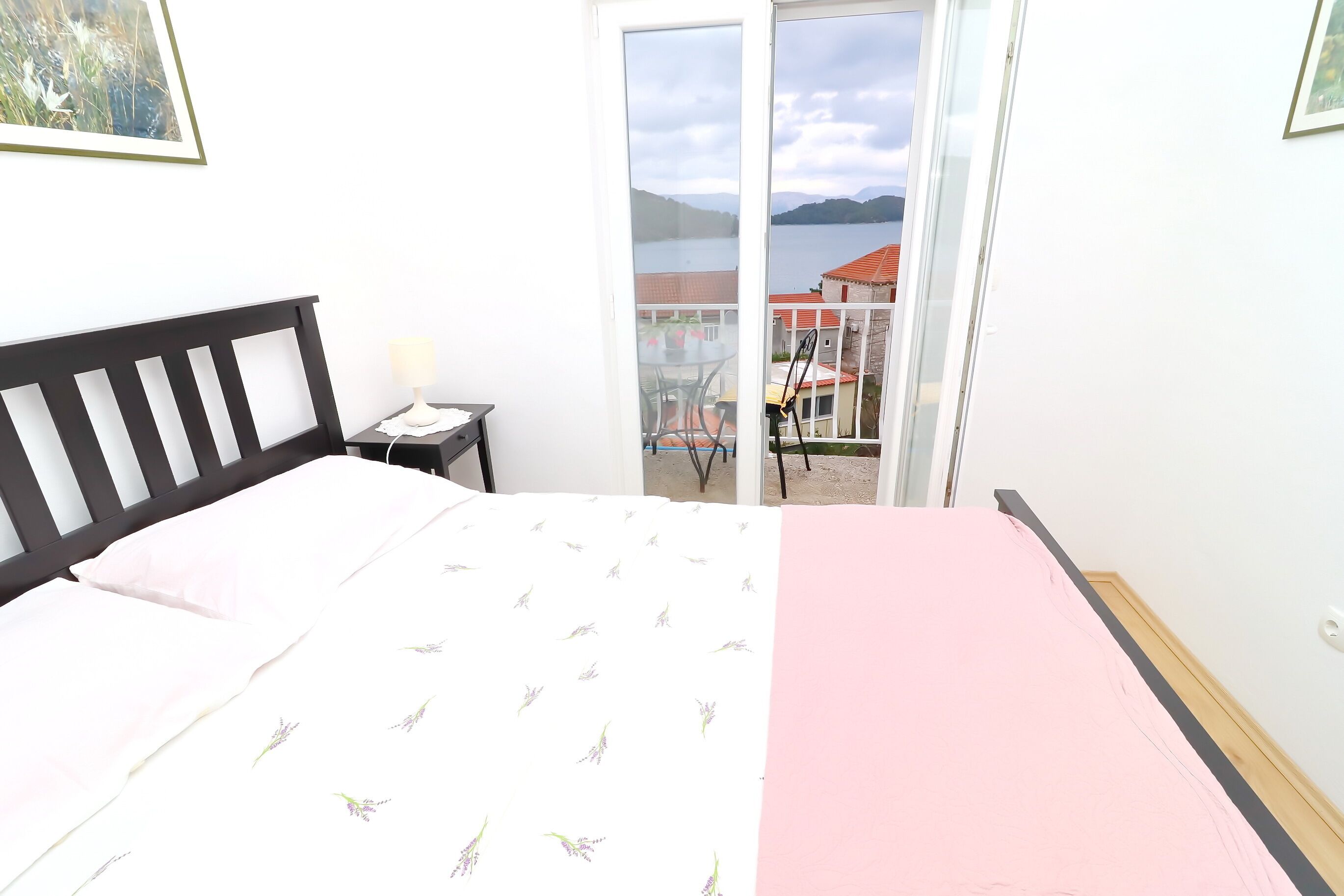 A bedroom with a bed, a nightstand, a lamp, and a view of a balcony with a table and chairs.