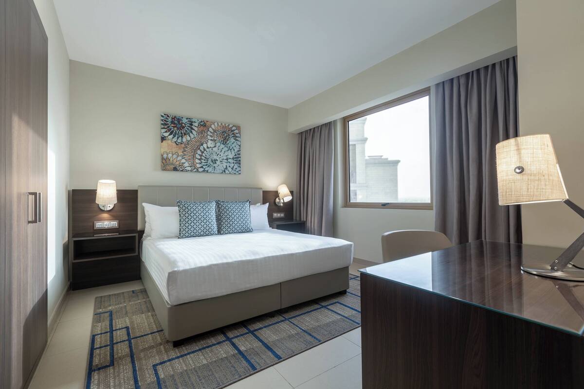 Une chambre d’hôtel moderne, dotée d’un grand lit, de deux tables de chevet avec des lampes, d’un bureau et d’une grande fenêtre avec des rideaux.