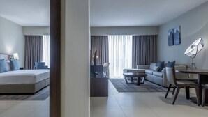 A modern hotel room with a bed, a sofa, a dining table, and a chair. There is a window with curtains, a floor lamp, and artwork on the wall. - Residence Inn by Marriott Algiers Bab Ezzouar (Algiers)