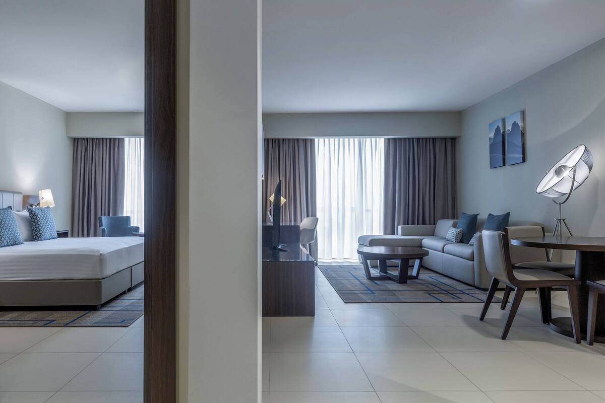 Une chambre d’hôtel moderne avec un lit, un canapé, une table à manger et une chaise. On y trouve une fenêtre avec des rideaux, une lampe de sol et des œuvres d’art accrochées au mur.