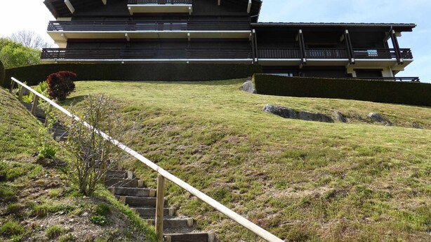 Een modern gebouw met een plat dak en meerdere balkons, gelegen op een grasachtige heuvel met een trap ernaartoe.