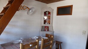 Een eetruimte met een tafel voor vier personen, houten stoelen, een klein bijzettafeltje, een wandplank met servies en een raam met jaloezieën.