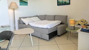 Ein modernes, kompaktes Zimmer mit einer grauen Schlafcouch, einem kleinen weißen Tisch, einer Stehlampe und zwei Bildern an den Wänden.