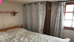 Un dormitori amb un llit, cortines, un reposacapses de fusta, una estant amb un objecte petit i una finestra.