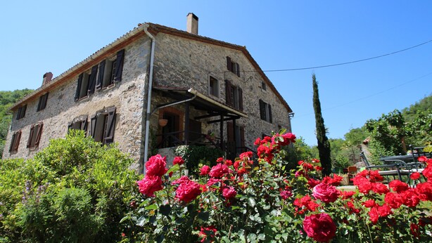 Un bâtiment en pierre doté d’un balcon, entouré d’une végétation luxuriante et de fleurs rouges.