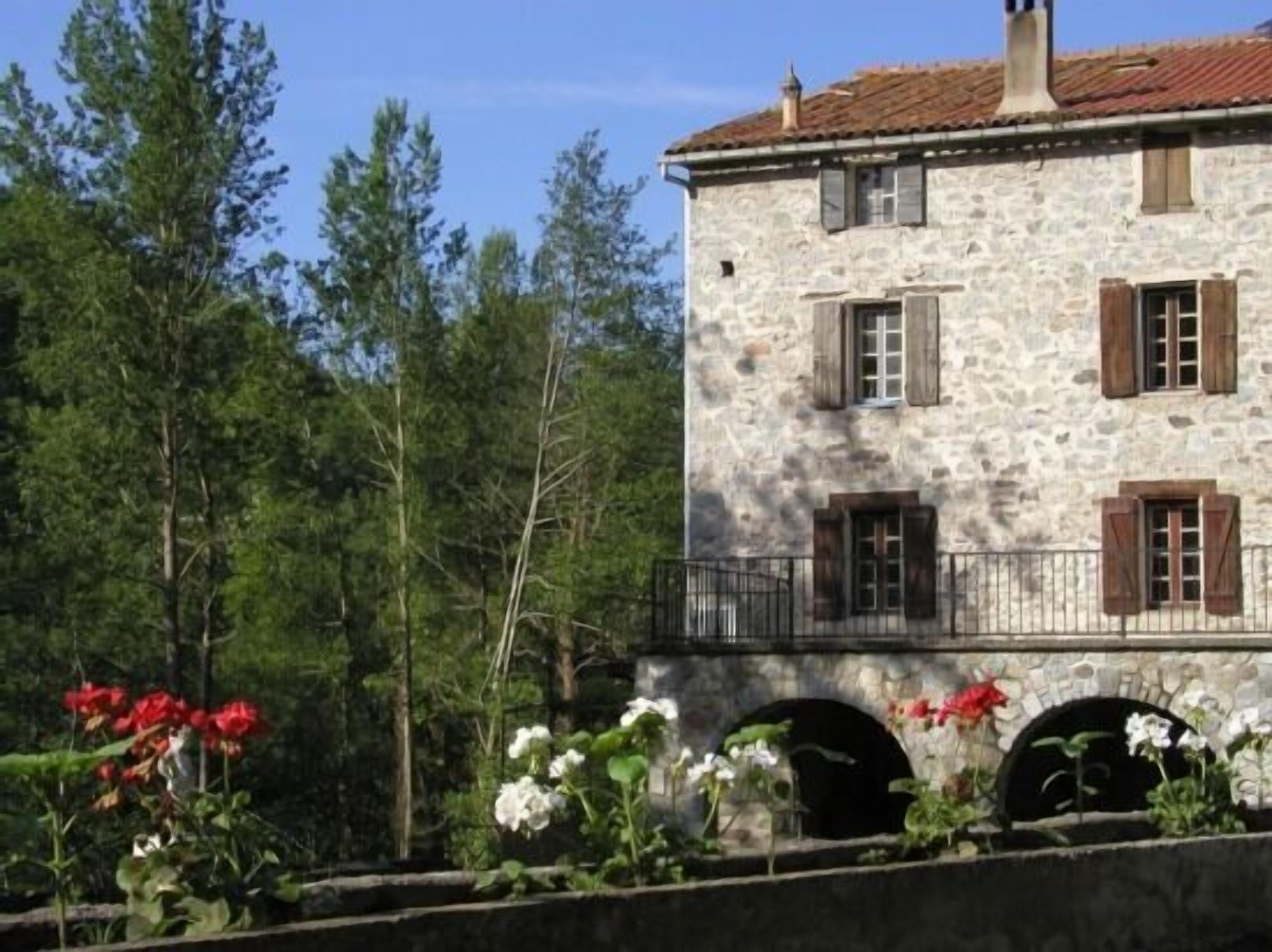 Un bâtiment en pierre doté de nombreuses fenêtres et volets, entouré de verdure et de fleurs.