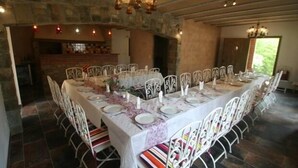 Une longue table à manger, entourée de chaises blanches, un lustre au-dessus et un mur de pierre en arrière-plan.