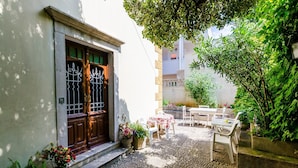 Un cortile con un edificio bianco, una porta in legno e un piccolo tavolo con sedie. Sono presenti piante in vaso e un pavimento in pietra.
