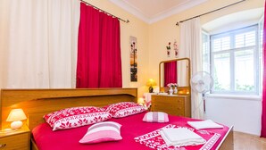 Camera d'albergo con un letto in legno, biancheria rossa, una lampada da comodino, uno specchio, una finestra con tende e una ventola.