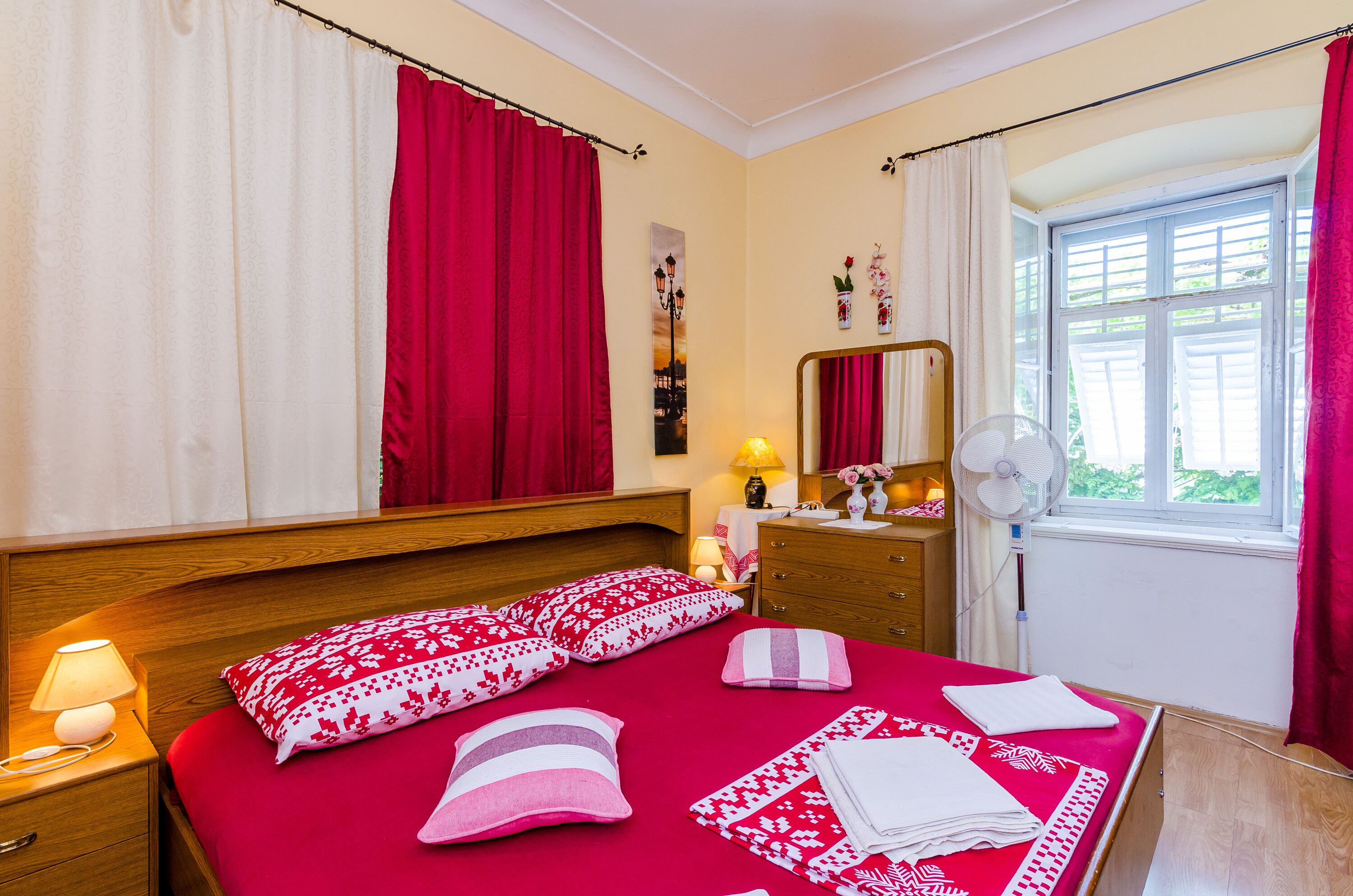 A bedroom with a wooden headboard, a bed with red bedding, a bedside table with a lamp, a dresser, a mirror, and a window with curtains.