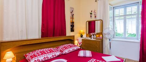 A bedroom with a wooden headboard, a bed with red bedding, a bedside table with a lamp, a dresser, a mirror, and a window with curtains.