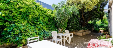 An outdoor patio with white tables and chairs, surrounded by lush greenery and a large tree.