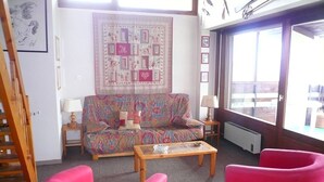 A cozy living room with a colorful sofa, a wooden coffee table, and bright pink chairs. A large window offers a view of the outdoors.