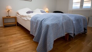 Un dormitorio con una cama, dos lámparas de noche, una mesita de noche de madera, una silla, una ventana con cortinas y un cuadro enmarcado en la pared.