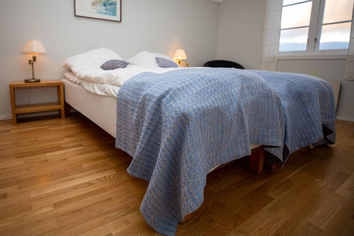 Ein Schlafzimmer mit einem Bett, zwei Nachttischlampen, einem Holznachttisch, einem Sessel, einem Fenster und einem Bild an der Wand.