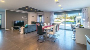 A modern living room with a flat-screen TV, a sofa, a dining table with chairs, and a kitchen area with bar stools. - Villa Dolores (Vrsar)