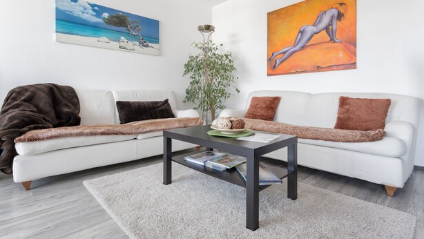 Ein modernes Wohnzimmer mit einem weißen Sofa, einem dunklen Couchtisch und einem großen Gemälde mit Strandmotiv.