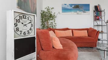 Ein Wohnzimmer mit einer großen Uhr, einem roten Sofa, einer Pflanze und einem Bücherregal.