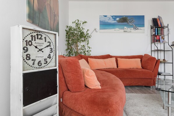 Un salon avec une grande horloge, un canapé rouge, une plante et une bibliothèque.