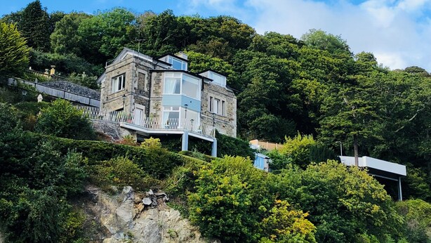 A modern house with a stone facade, situated on a hillside with lush greenery and a clear blue sky. - EXCLUSIVE HOUSE PRIVATE BEACH SEA FRONT SLEEPS 13 AMAZING VIEWS (Menai Bridge)