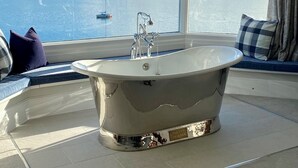 A modern bathroom with a freestanding bathtub, large windows offering a view of a lake and mountains, and a cozy seating area with cushions. - EXCLUSIVE HOUSE PRIVATE BEACH SEA FRONT SLEEPS 13 AMAZING VIEWS (Menai Bridge)