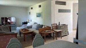 Un salón con un sofá, una mesa de centro y un televisor. Hay dos sillones y una mesita con una lámpara. Las paredes son de tonos claros y hay una puerta que conduce a otra habitación.