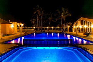 A nighttime swimming pool with illuminated blue lighting, surrounded by palm trees and buildings with warm lighting.