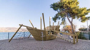 Una estructura de juegos de barco pirata de madera con una red de cuerda, ubicada en una zona de grava con vistas al mar y colinas al fondo.