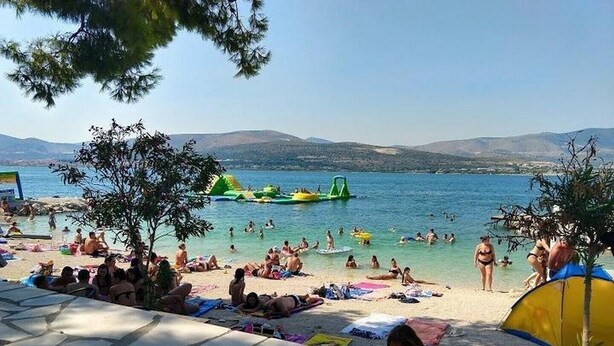 Una playa con gente tomando el sol y nadando, un área de piscina y vistas al mar y colinas al fondo.