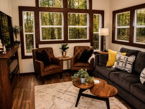 A cozy living room with a brown leather sofa, two brown leather armchairs, a wooden coffee table, and a small side table with a plant. Large windows with dark frames offer a view of trees. - Adventurer at The Retreat at Water's Edge (Tracy City)
