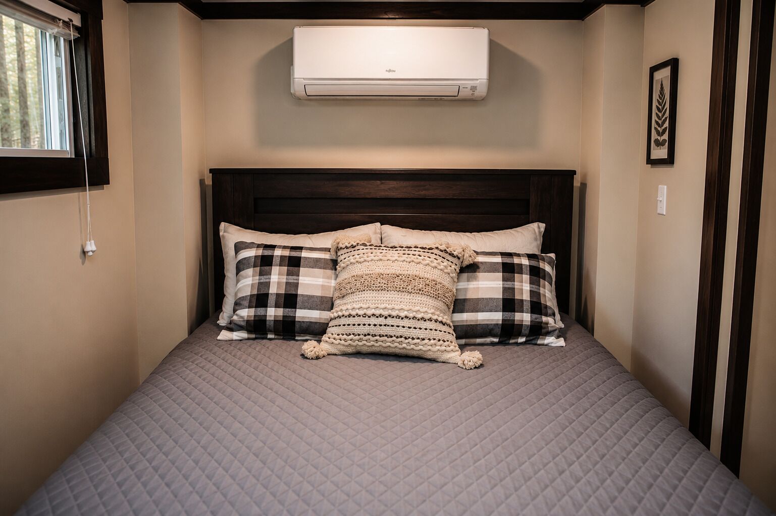 A bed with a quilted cover, two pillows, a wall-mounted air conditioner, and a window with blinds.