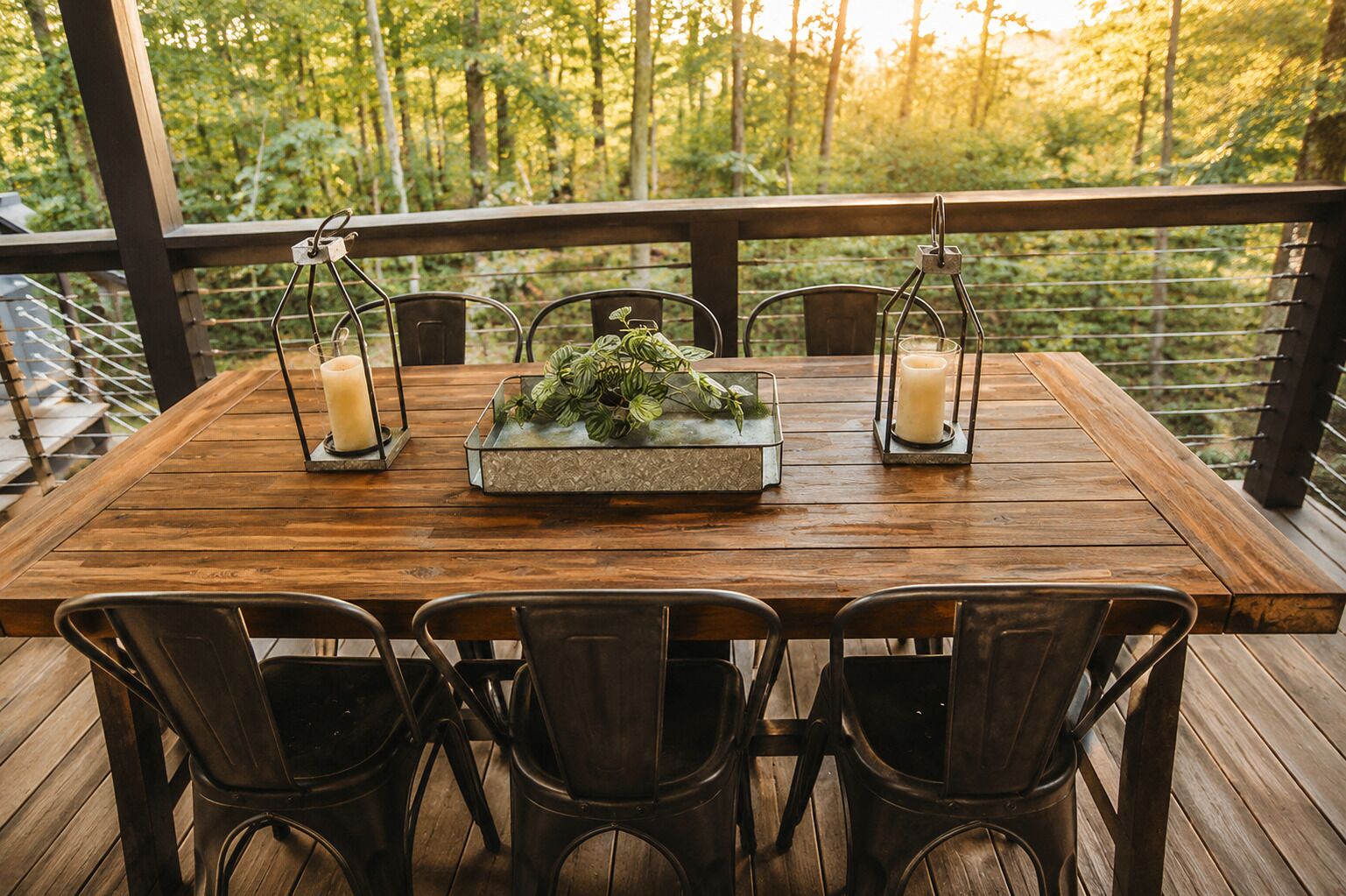 A wooden deck with a table set for four, surrounded by metal chairs, with candles and a plant centerpiece.