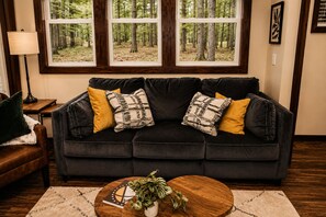 Ein dunkelbraunes Sofa mit gemusterten Kissen, ein runder Holztisch mit einer Pflanze und Blick auf einen Wald durch große Fenster.