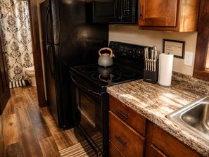 A compact kitchen with a black refrigerator, a stove, and a microwave. There is a wooden cabinet with a sink and a countertop. - Adventurer at The Retreat at Water's Edge (Tracy City)