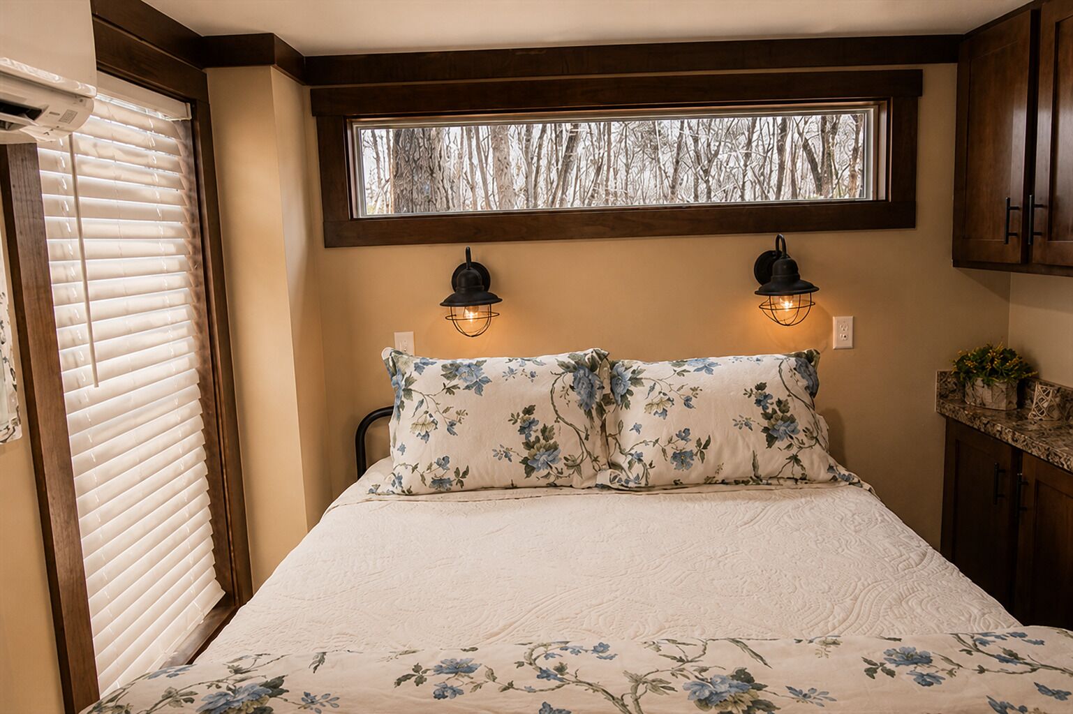 A bedroom with a bed, two wall-mounted lamps, a window with a view of trees, and a wooden headboard.