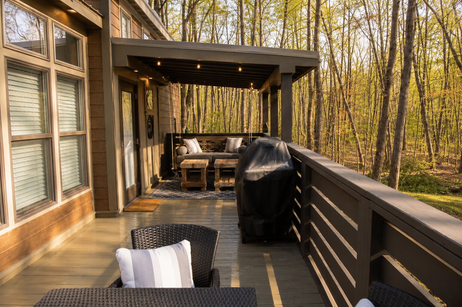 A covered outdoor deck with seating, a grill, and a view of a forest.