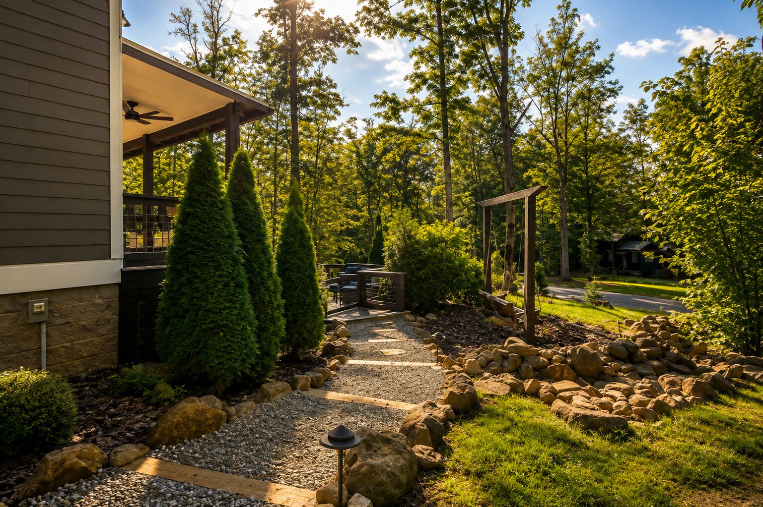 A pathway leading to a covered outdoor seating area with a pergola, surrounded by a landscaped garden with rocks and trees.
