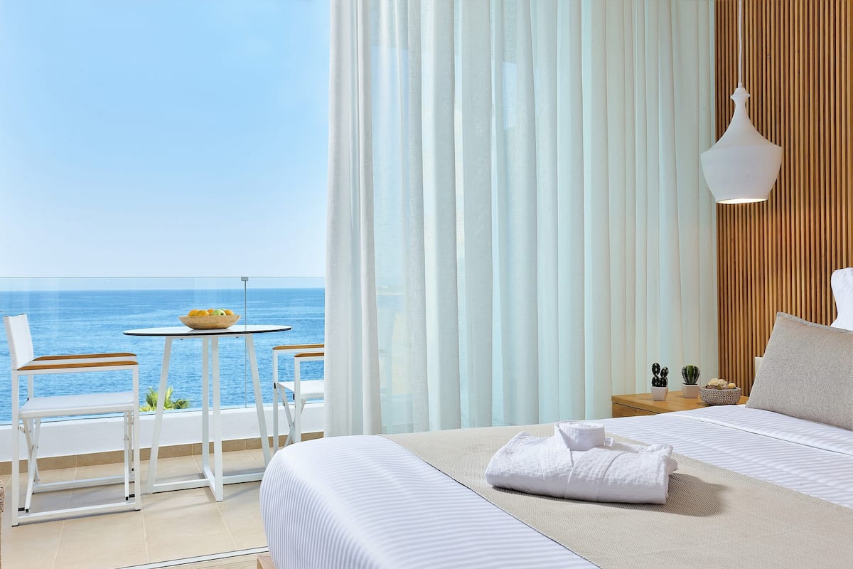 A hotel room with a bed, a small table with a bowl of fruit, two chairs, and a view of the sea.