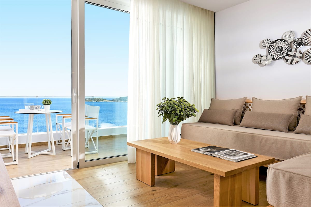 A modern living room with a wooden coffee table, a beige sofa, and a white dining table with chairs. A large window offers a view of the sea.