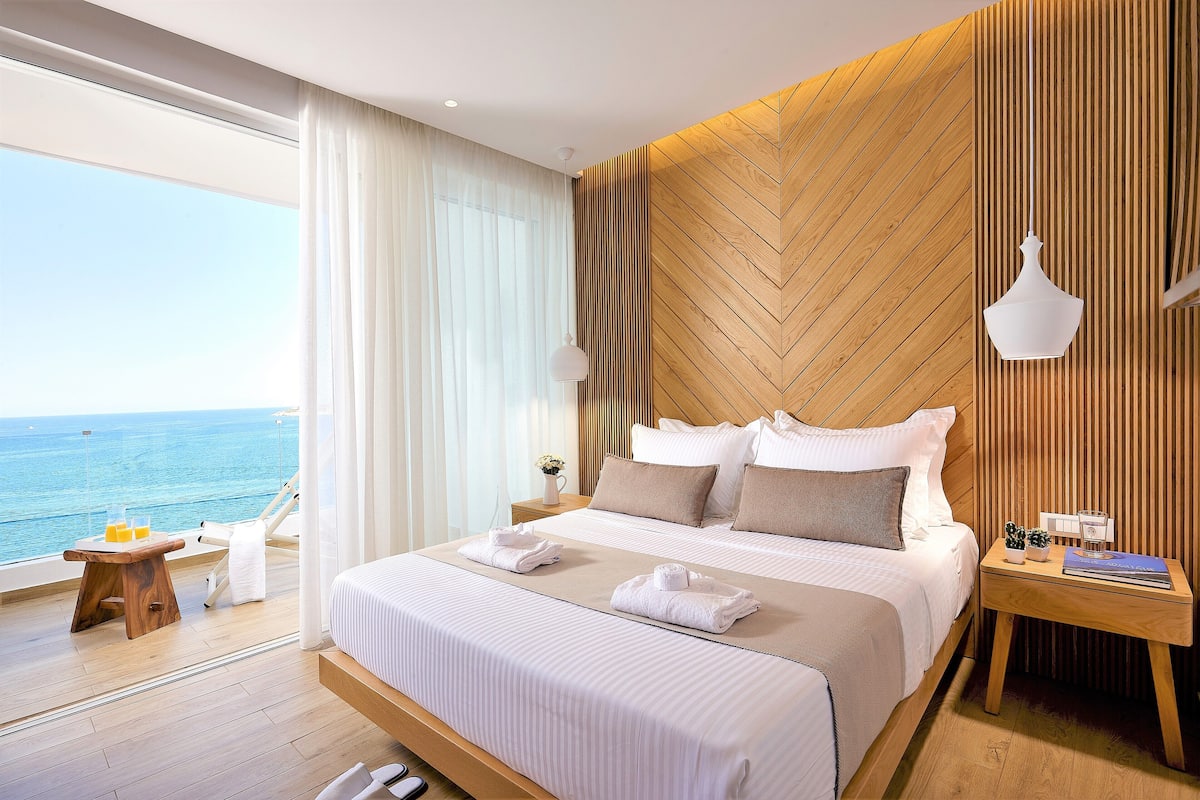 A modern hotel room with a large bed, wooden panel wall, and a view of the sea.