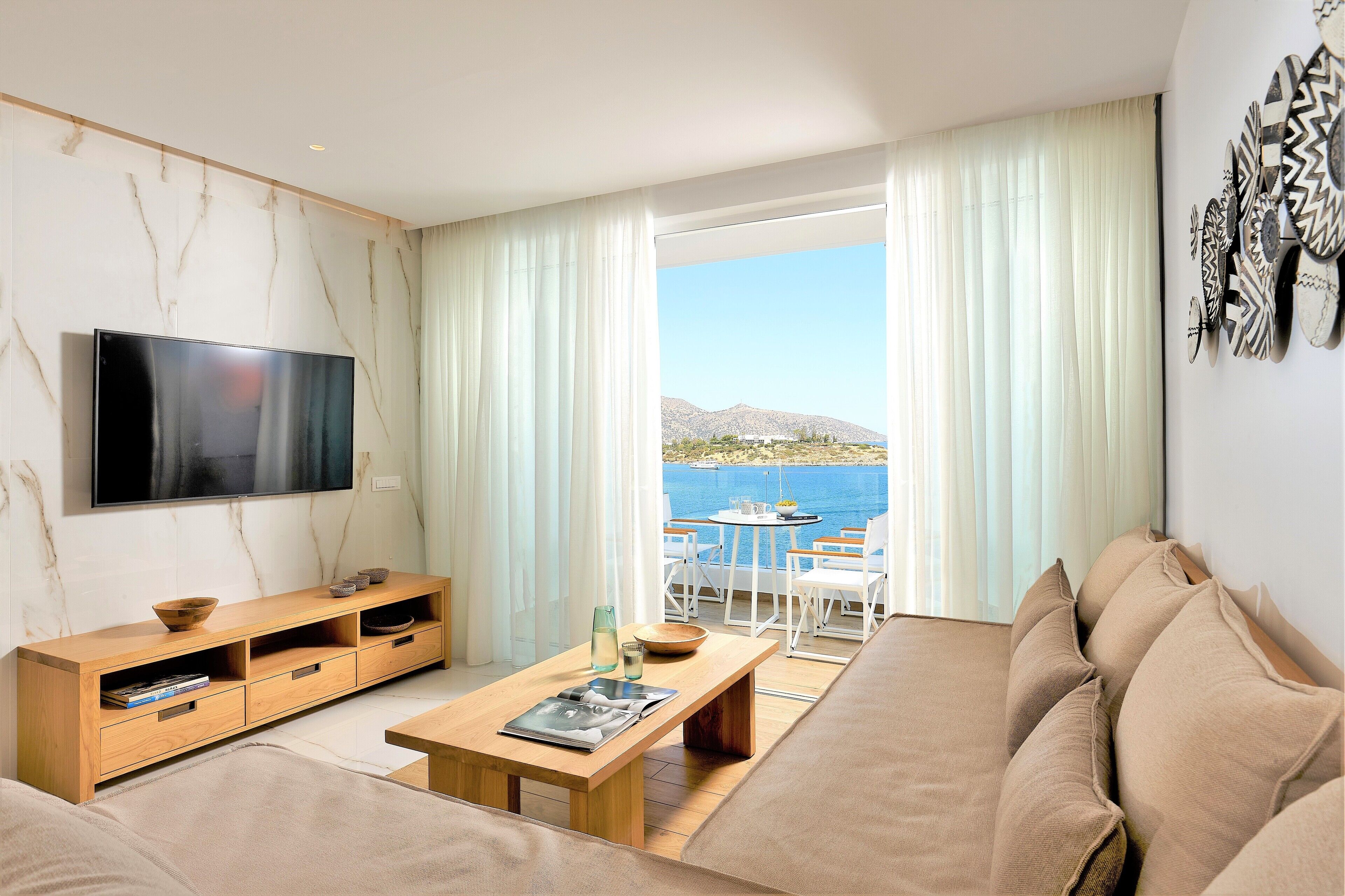 Una sala moderna con televisor de pantalla plana, mueble de madera para entretenimiento y un sofá. Se ve un comedor con una mesa y sillas a través de un ventanal con vistas a una masa de agua.