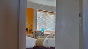 Ein Flur, der zu einem Schlafzimmer mit Bett, Fenster mit Jalousien und einer Pendelleuchte führt.