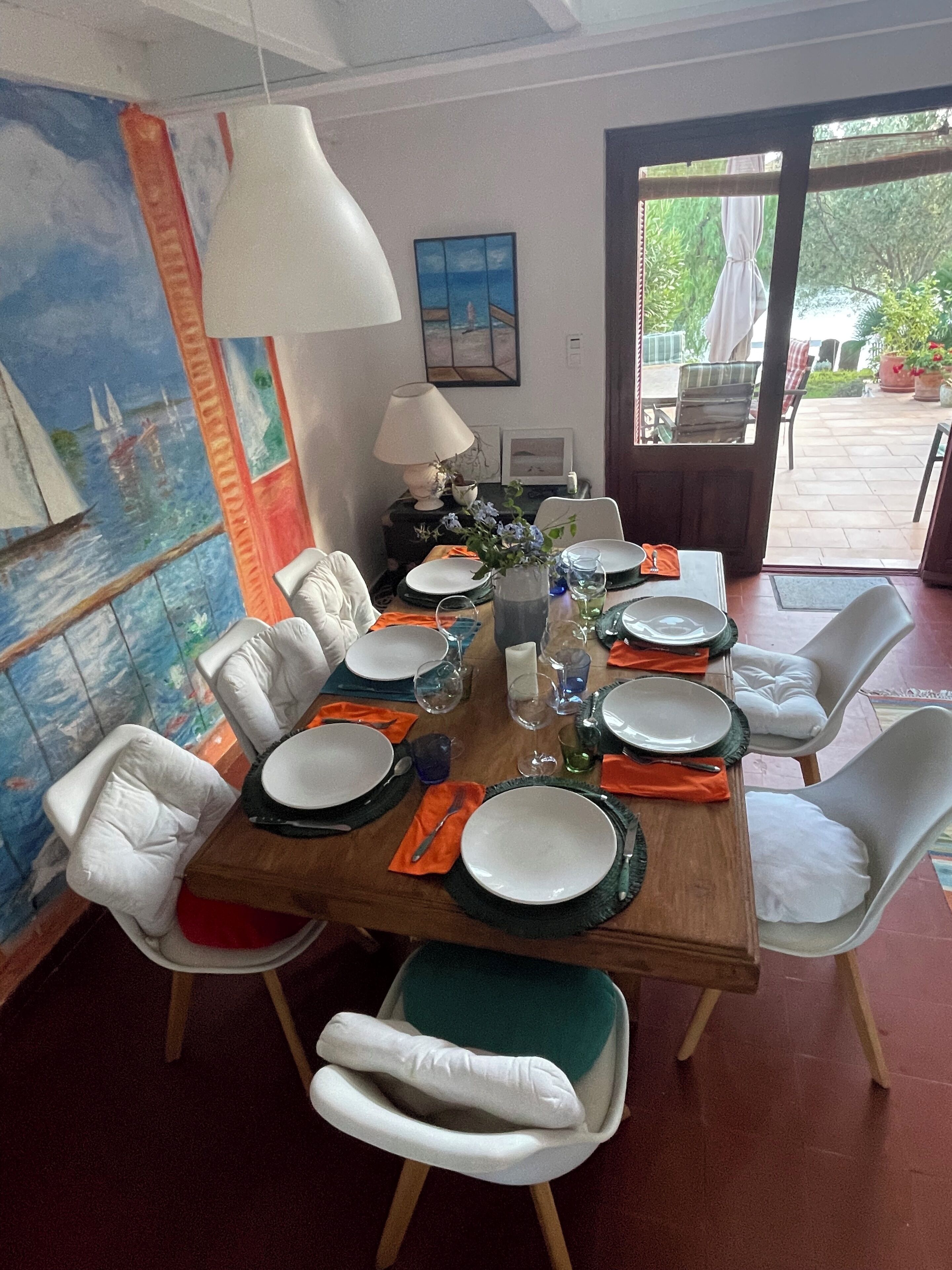 A dining area with a wooden table set for six, white cushioned chairs, a painting on the wall, and a view through the door.